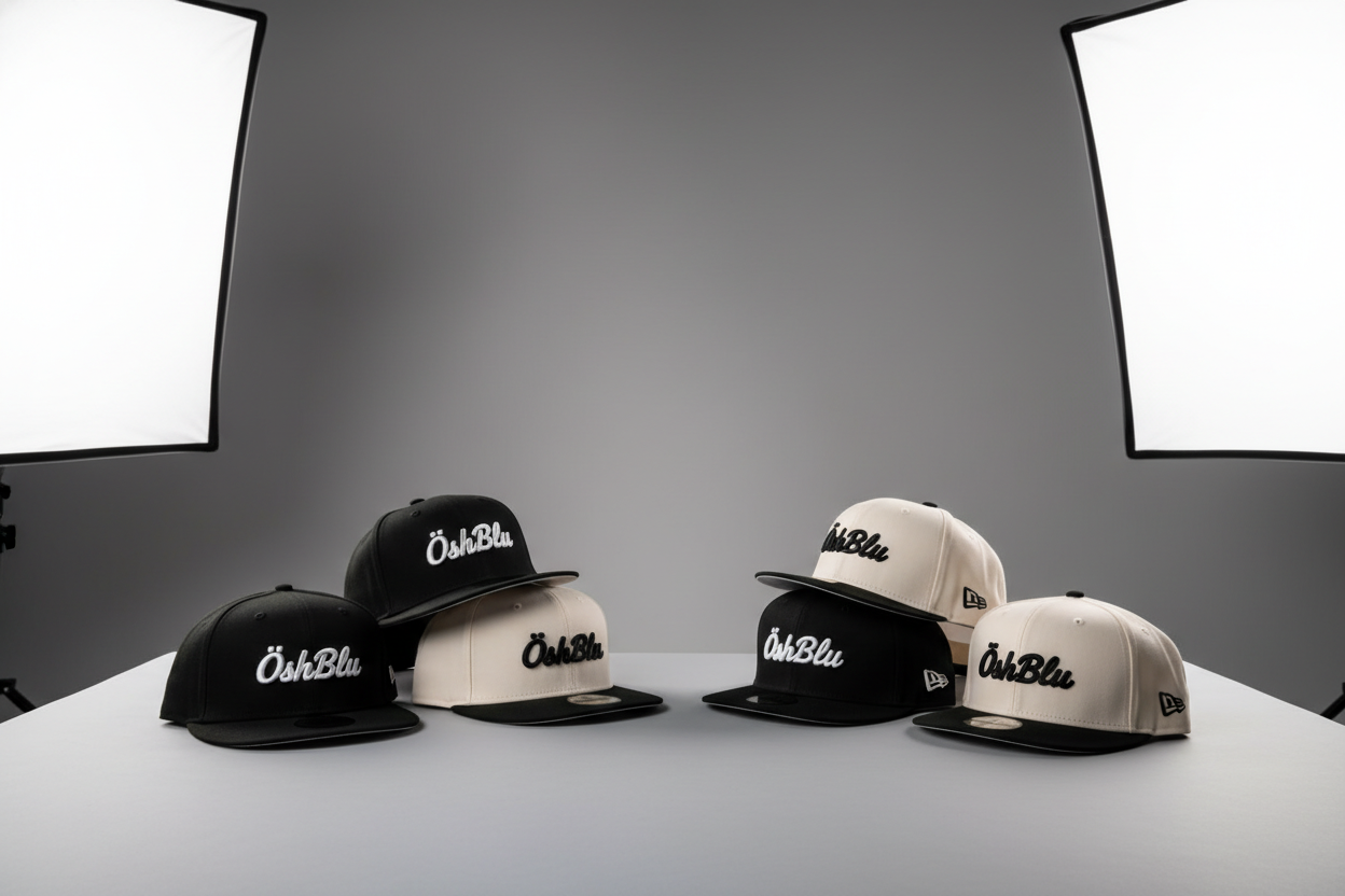 Estudio fotográfico minimalista, fondo neutro en tonos gris suave. Iluminación suave tipo softbox creando sombras delicadas y definidas. Colocar varias gorras de la marca ÖshBlu en primer plano sobre una superficie limpia tipo mesa de estudio. Modelos de gorras estilo New Era 9Fifty y 59Fifty. Incluir dos combinaciones de colores: (1) gorra negra con detalles blancos, logo ÖshBlu al frente en bordado blanco, visera negra; (2) gorra blanco hueso con visera negra, logo ÖshBlu en negro. Composición elegante y 